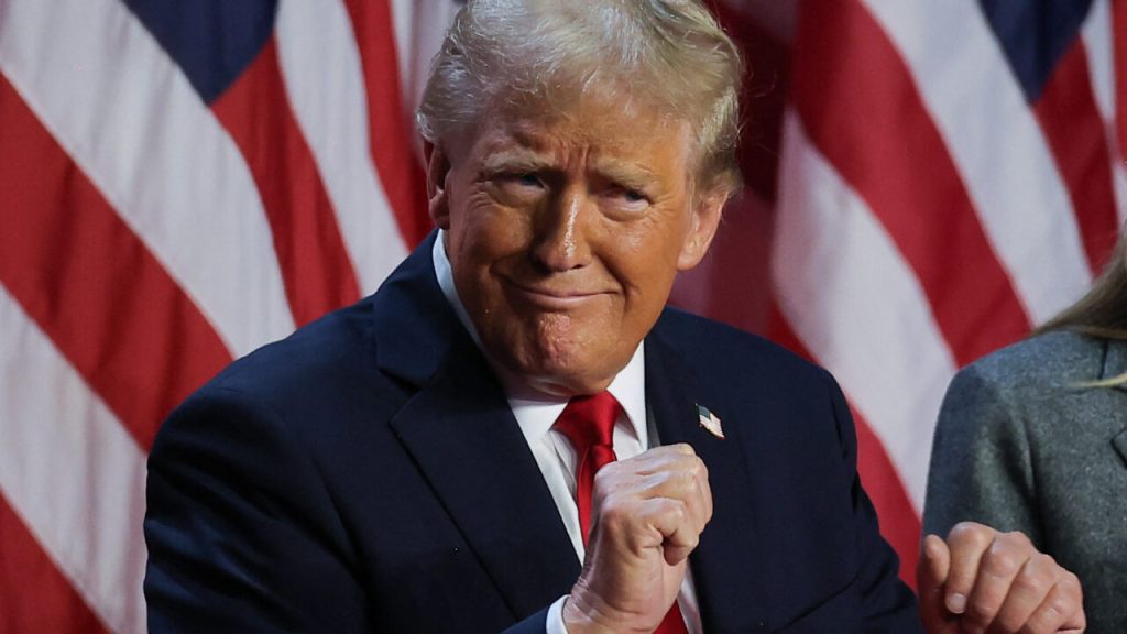 Donald Trump in a dark suit and red tie, posing in front of American flags with a confident expression.