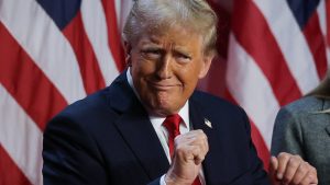 Donald Trump in a dark suit and red tie, posing in front of American flags with a confident expression.